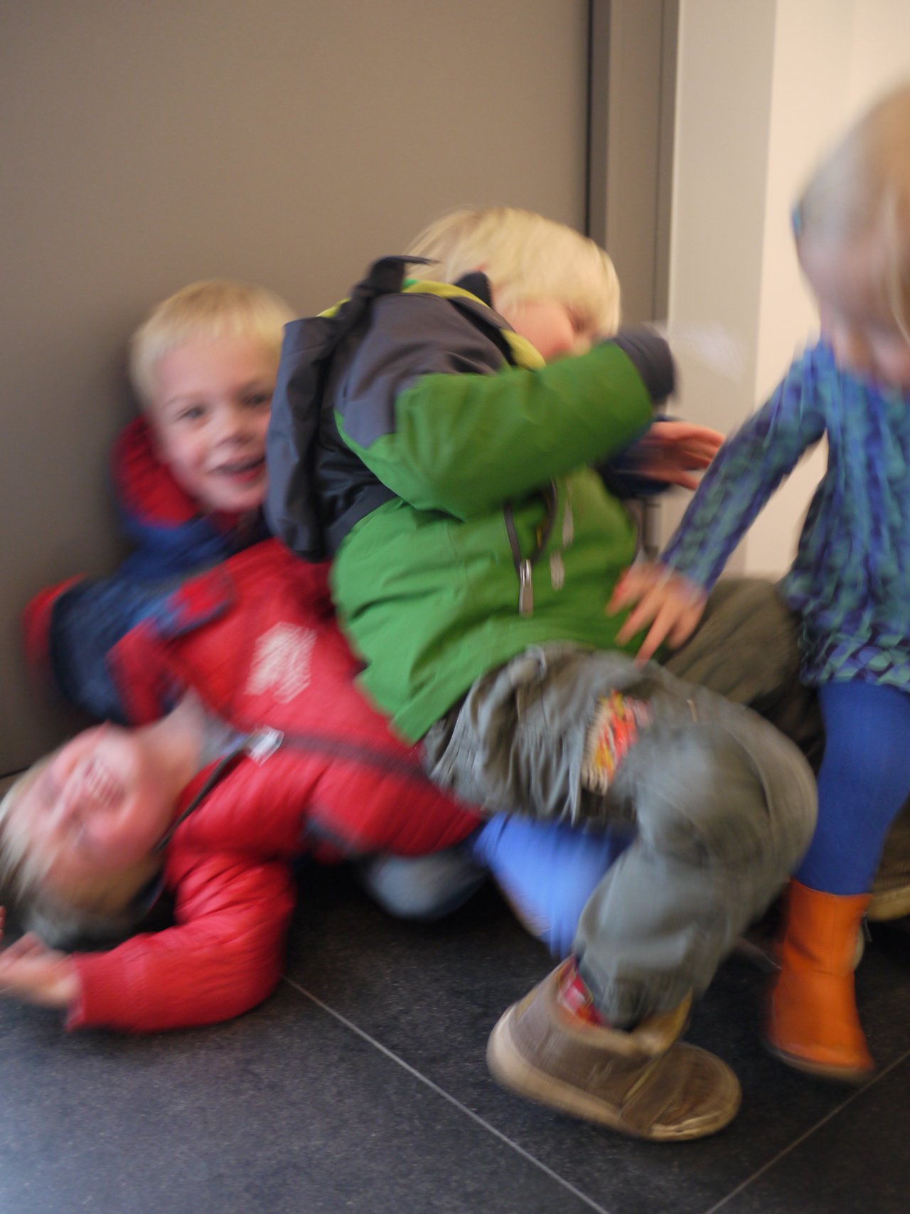 Several children in colorful winter clothing are playfully tumbling and piling on top of each other near a door.