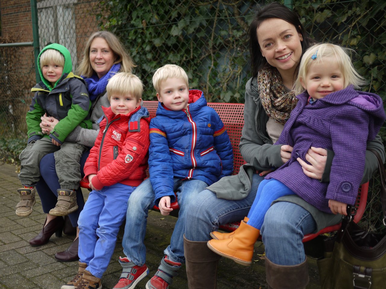 Two women sit on a bench holding young children, all dressed in warm clothing and smiling at the camera.