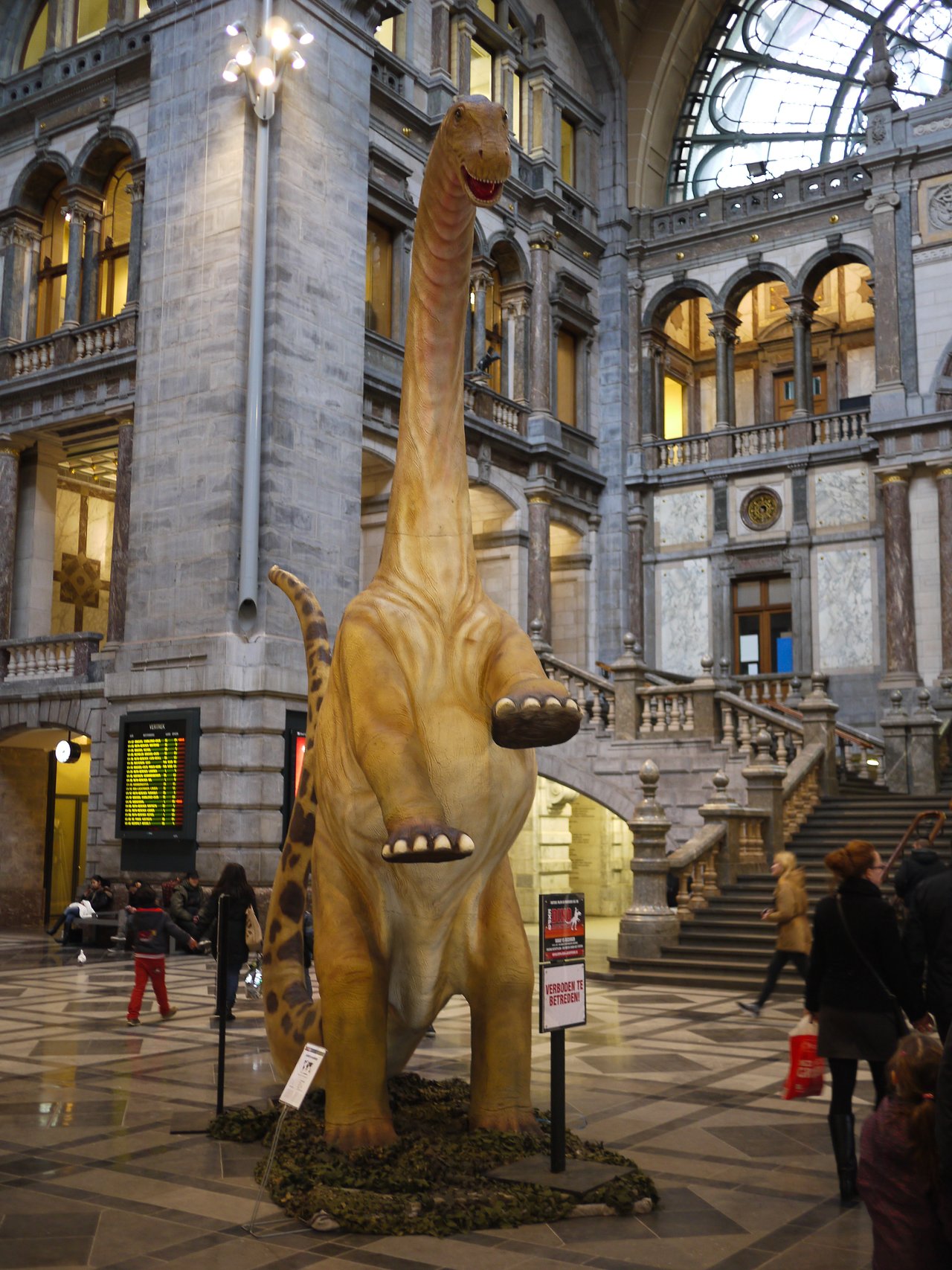 A large dinosaur statue stands in the middle of a grand indoor space with people walking around.