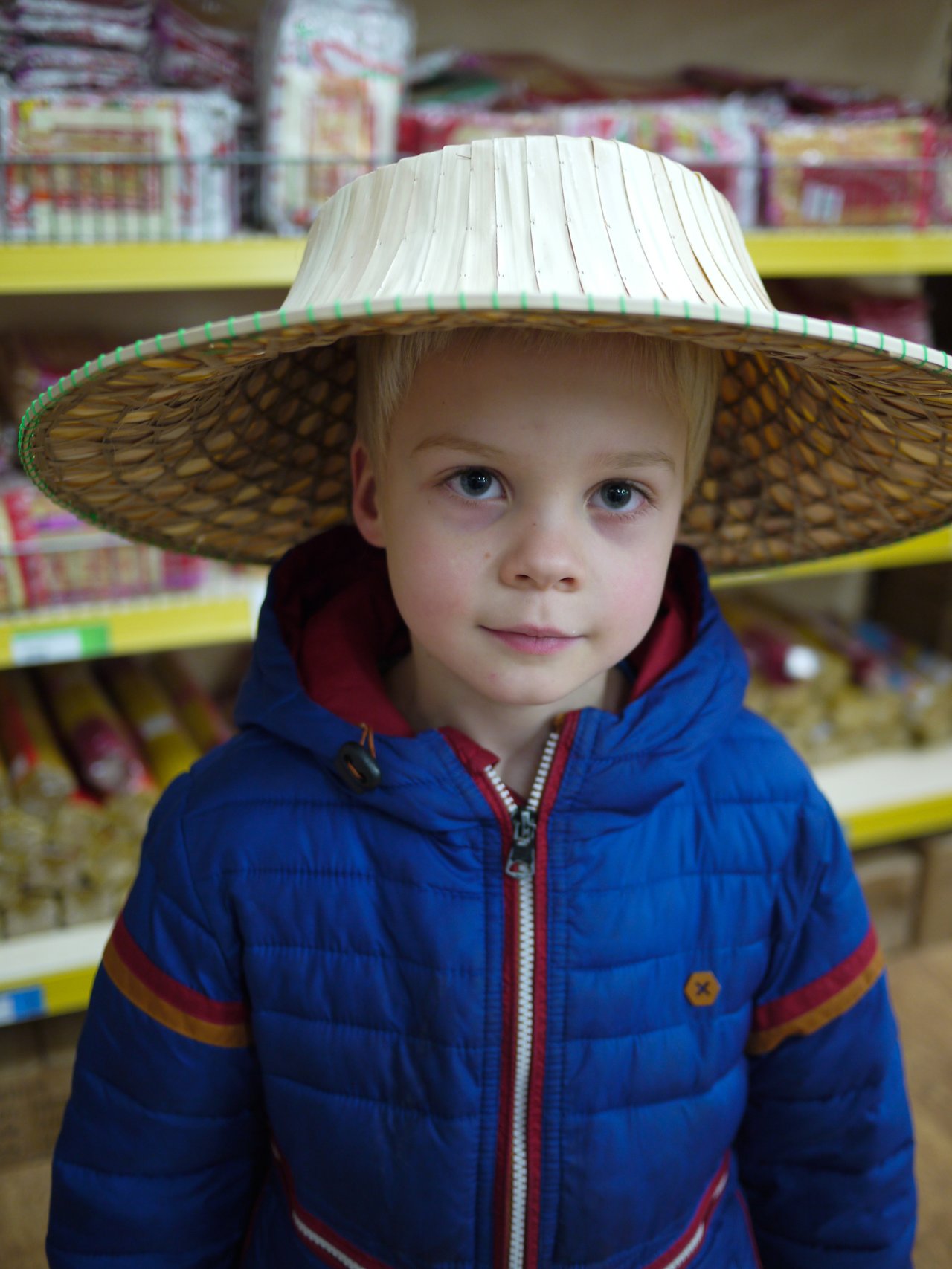 A child in a blue jacket wears a wide-brimmed straw hat inside a Chinese supermarket.