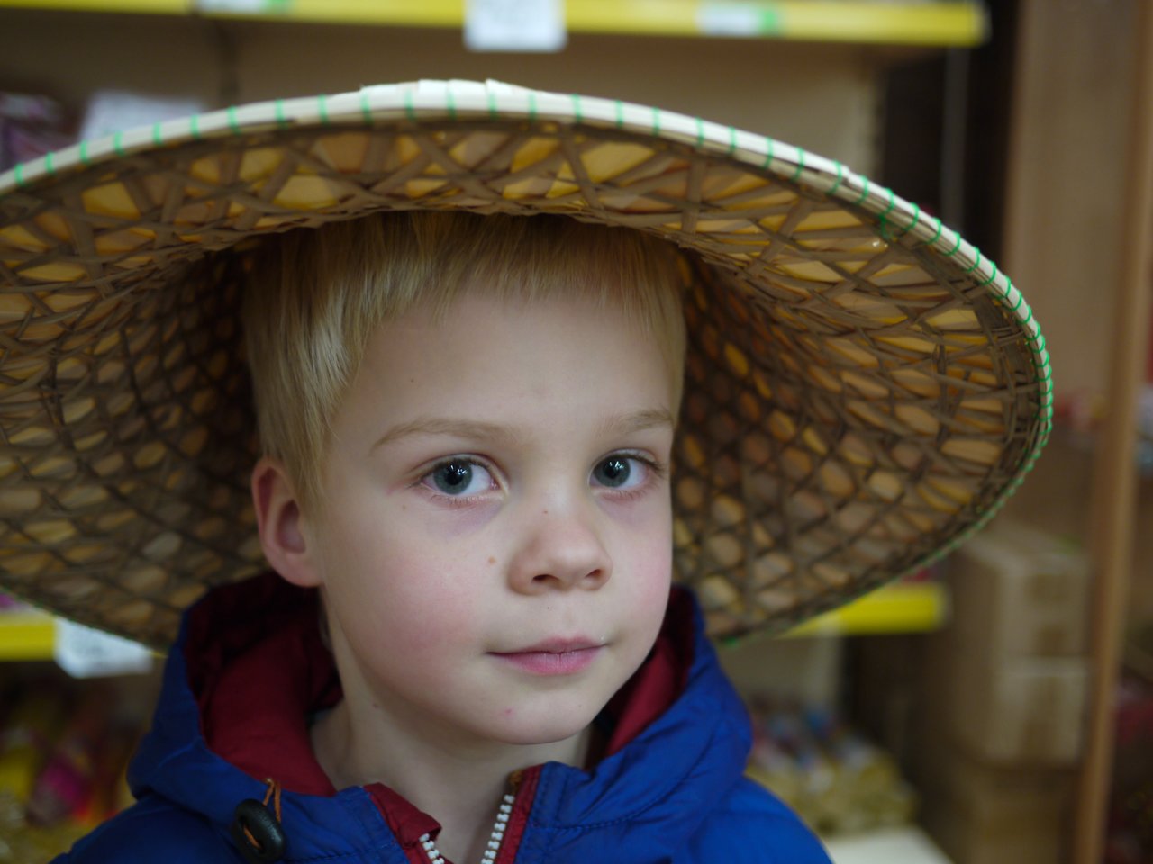 A child in a blue jacket wears a large woven hat inside a supermarket, looking directly at the camera.