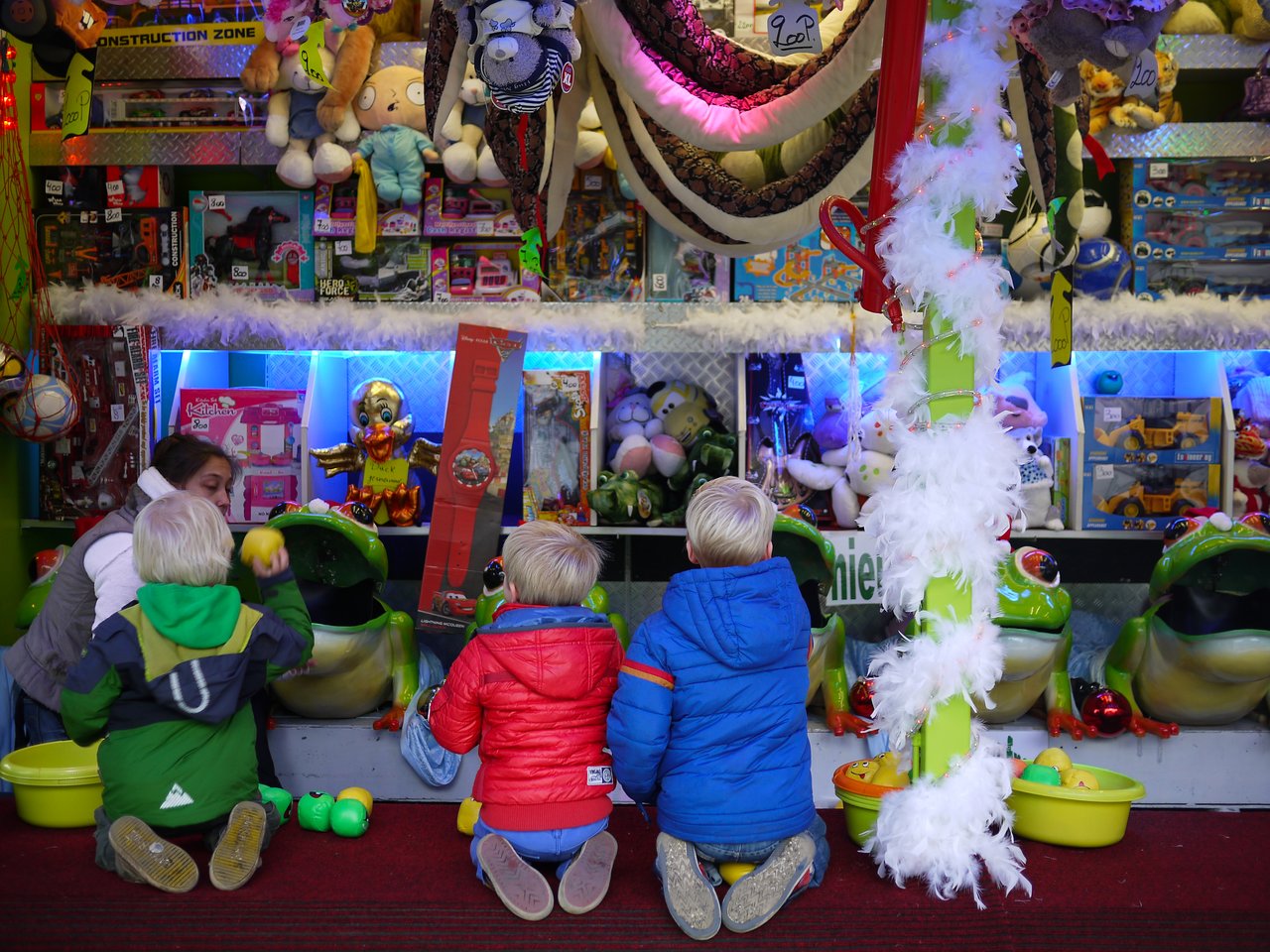 Three children play a ball-toss game at a carnival booth filled with toys and stuffed animals.