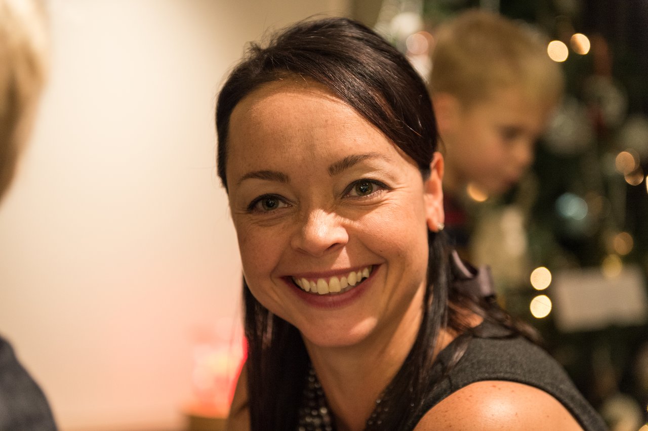 A woman smiles at the camera with a Christmas tree and a child in the background.