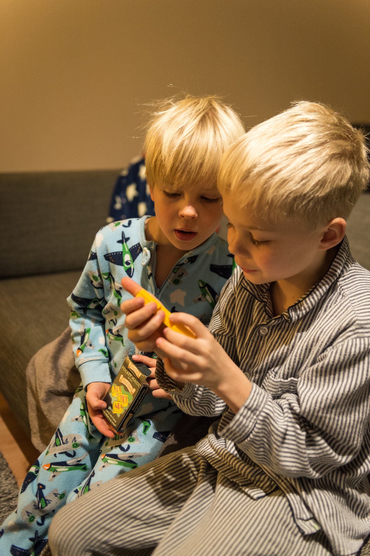Two young children in pajamas sit together, examining a small yellow object while holding a box of game cards.