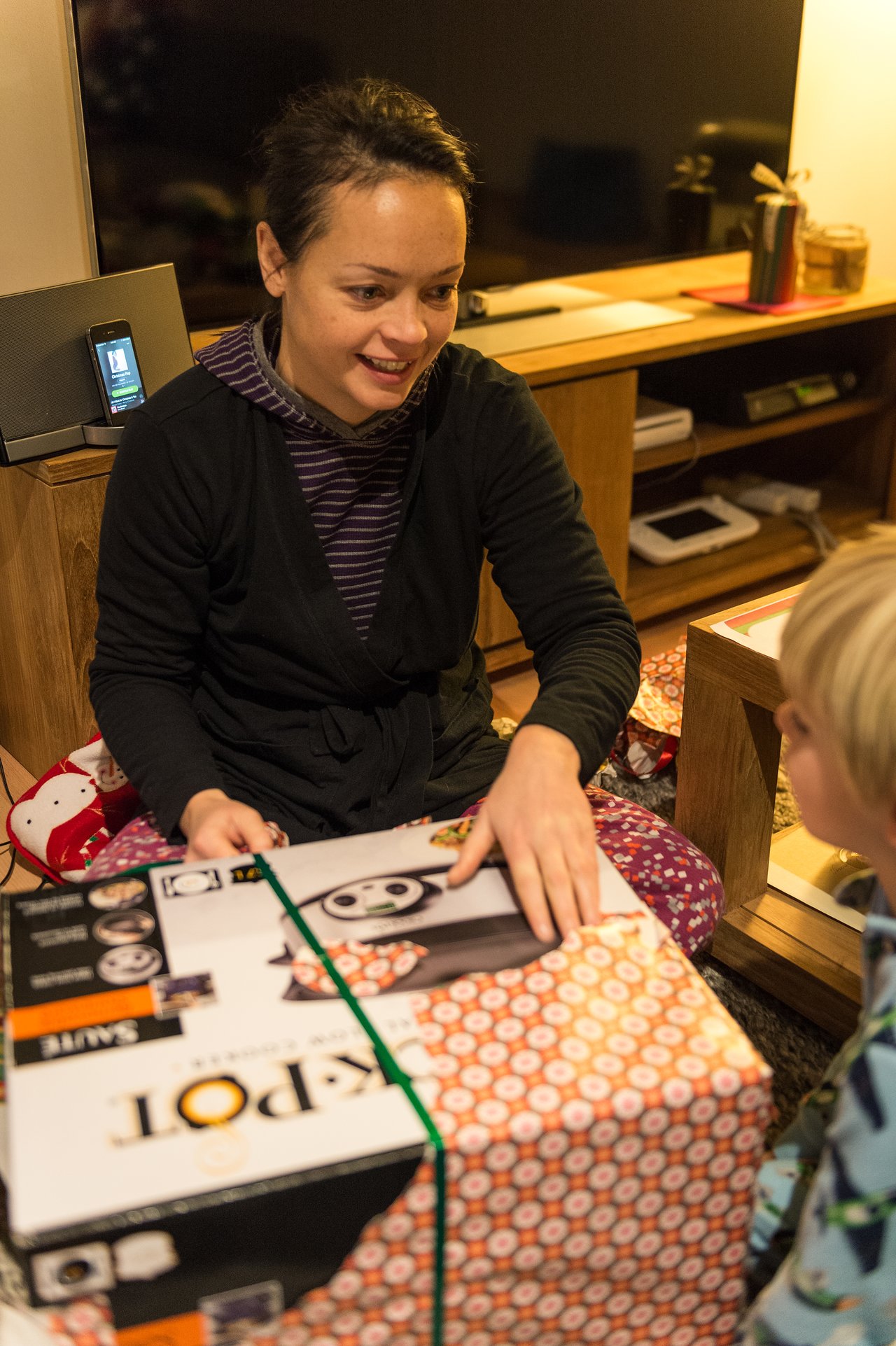 A woman sits on the floor, smiling while unwrapping a large Christmas gift, as a child watches.
