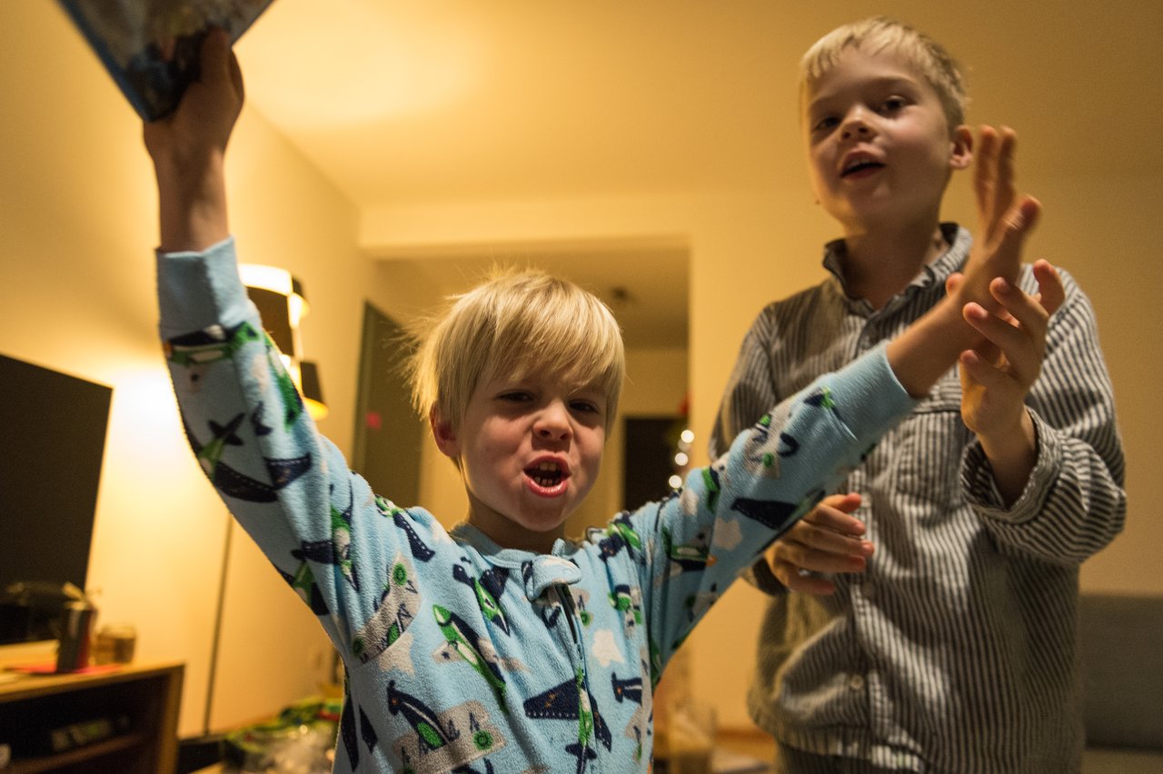 A young child in pajamas excitedly raises a gift while another child stands nearby, smiling.