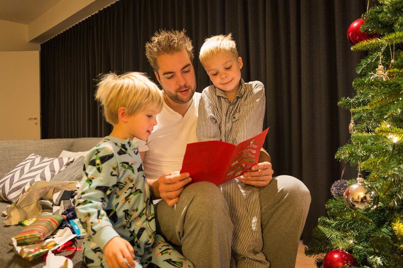 A man and two children sit by a Christmas tree, reading a red card together.
