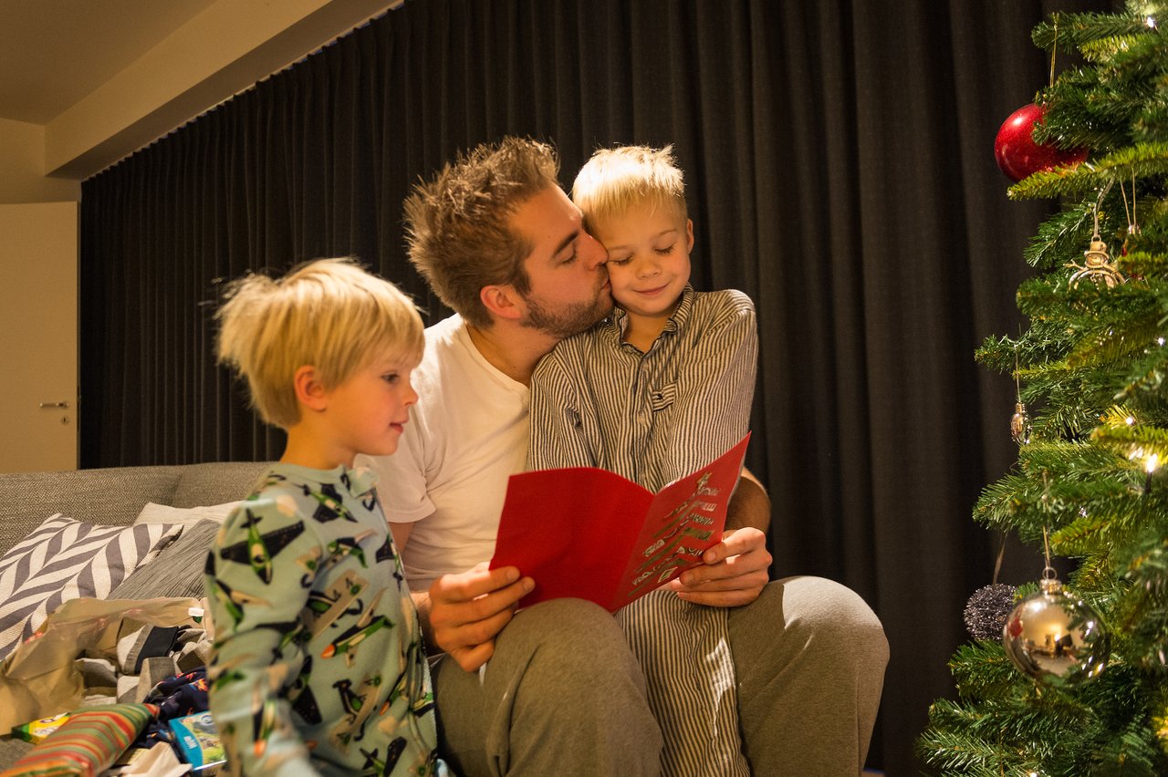 A father reads a Christmas card to his two young children near a decorated Christmas tree.