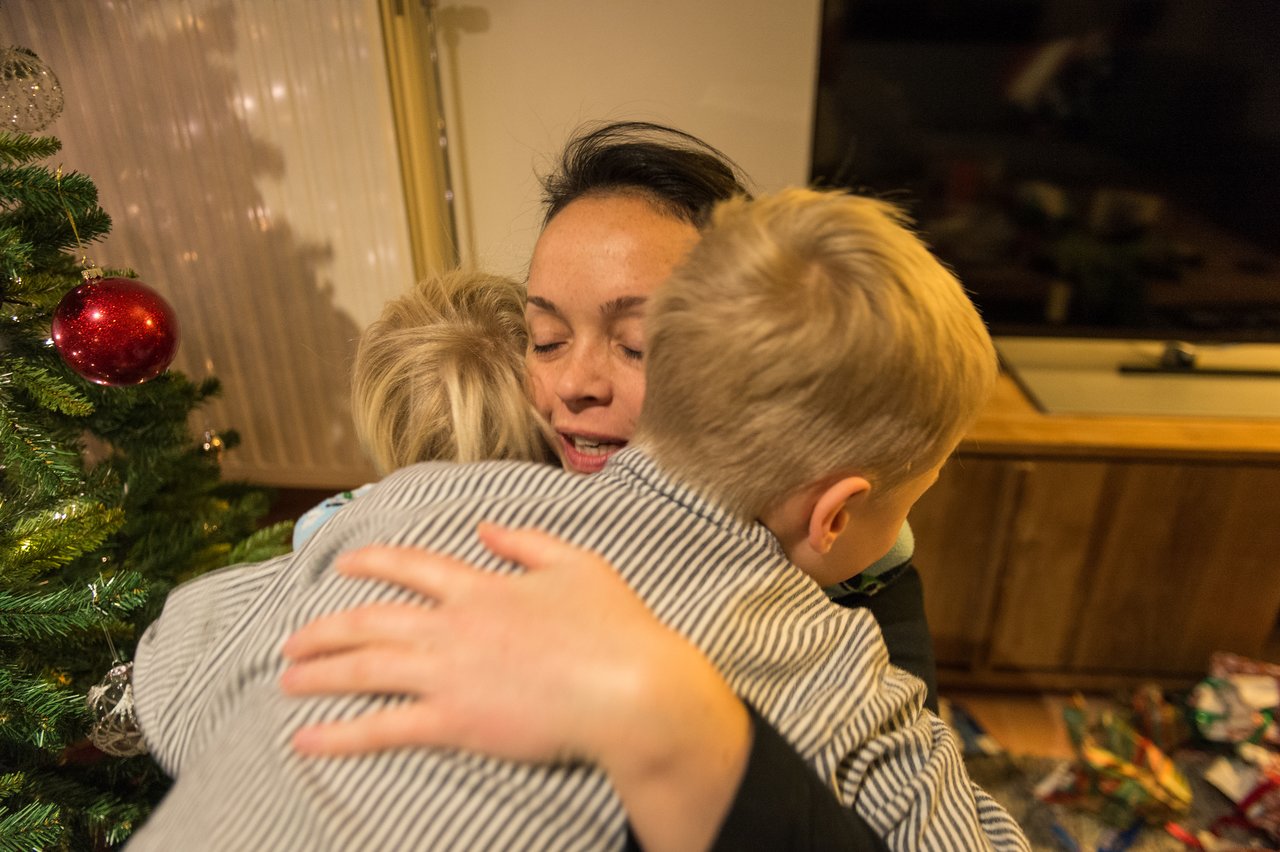 A woman hugs two children near a Christmas tree, with wrapped gifts on the floor in the background.
