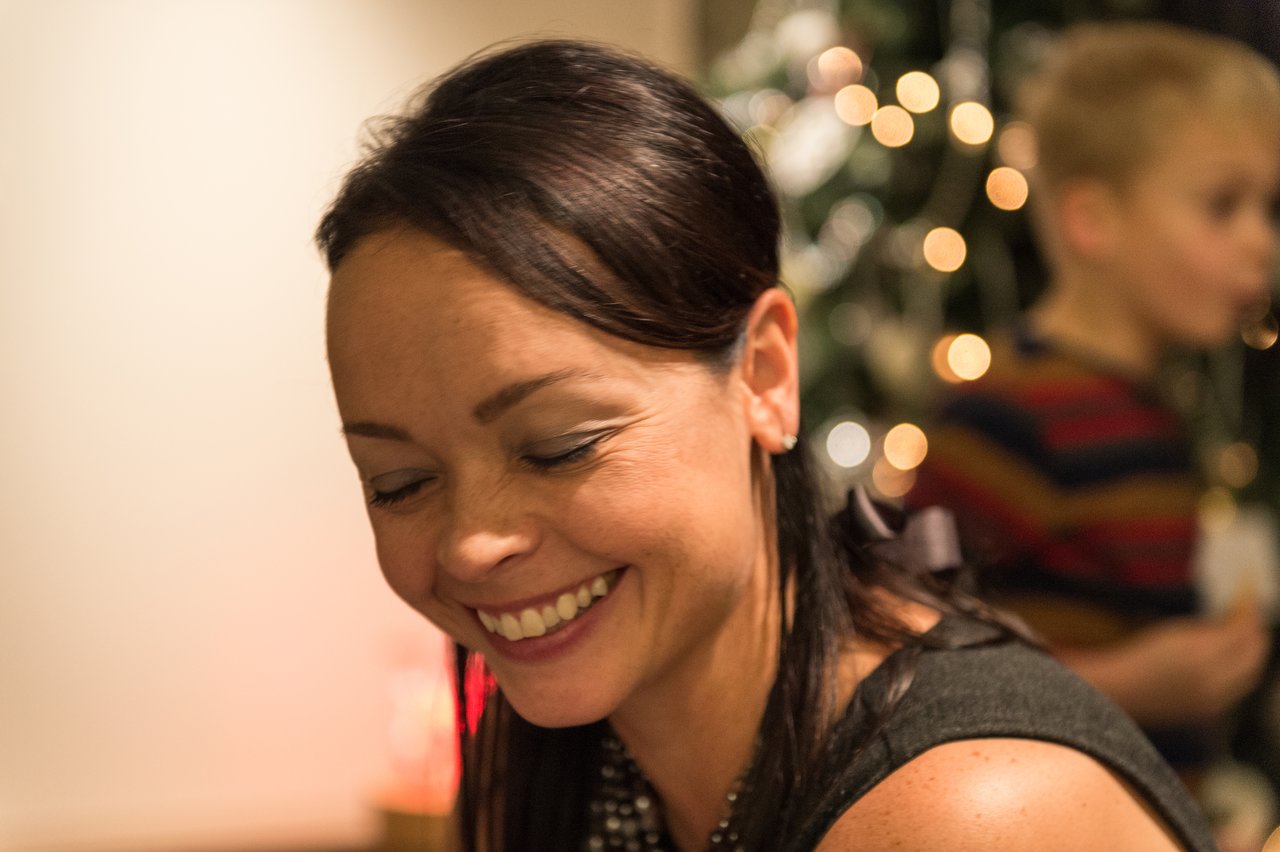 A woman smiles with her eyes closed, while a child in the background stands near a decorated Christmas tree.