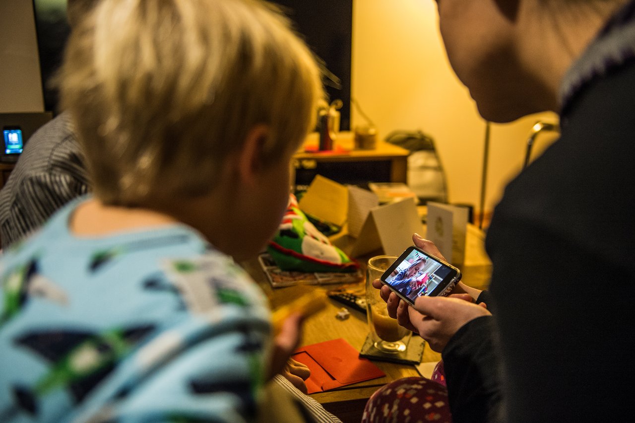 A child in pajamas watches as an adult shows a photo on a smartphone in a cozy setting.