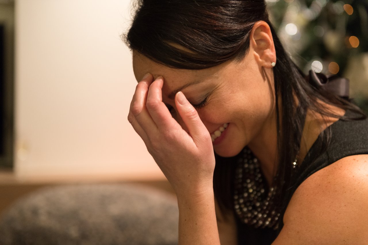 A woman in a black dress smiles and covers her face with her hand, appearing joyful.