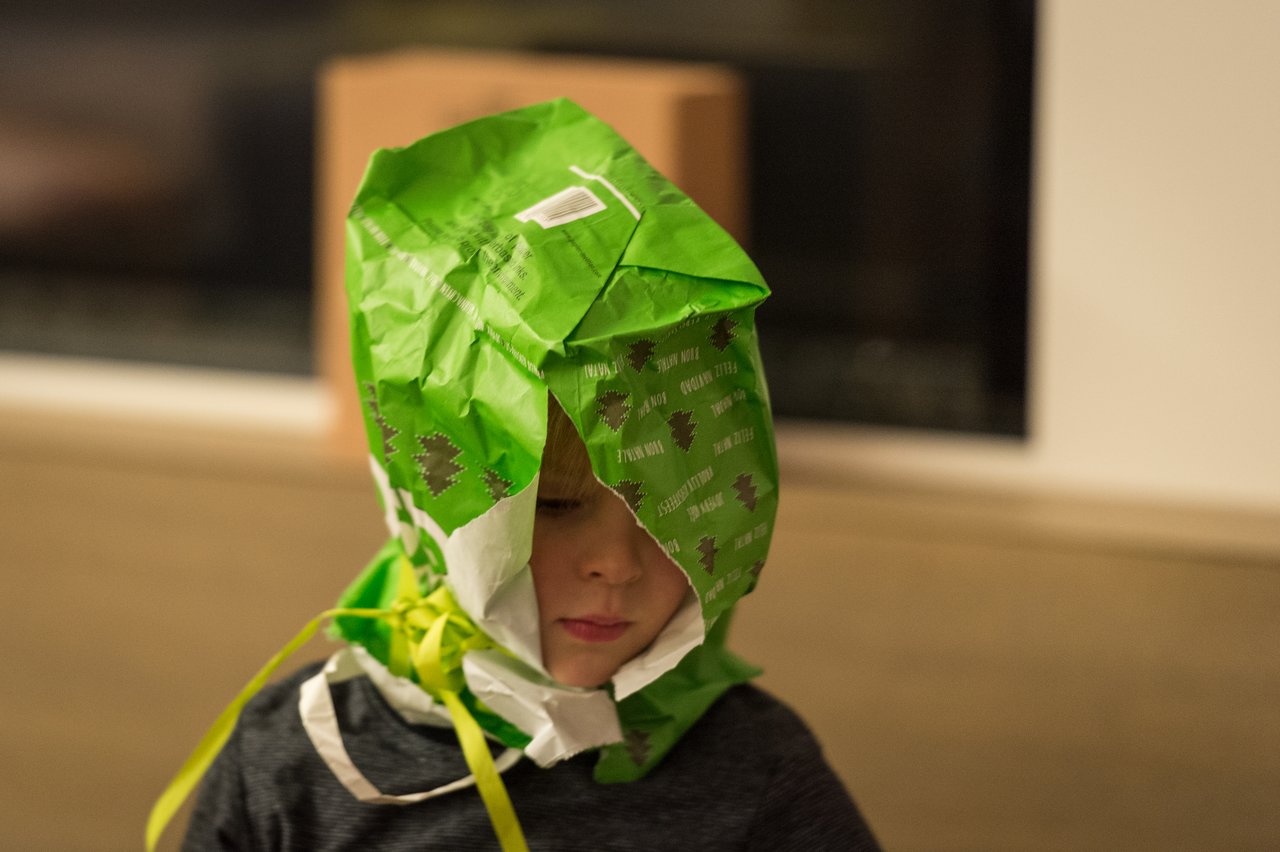 A child wears a torn green gift bag on their head, with ribbons hanging down.