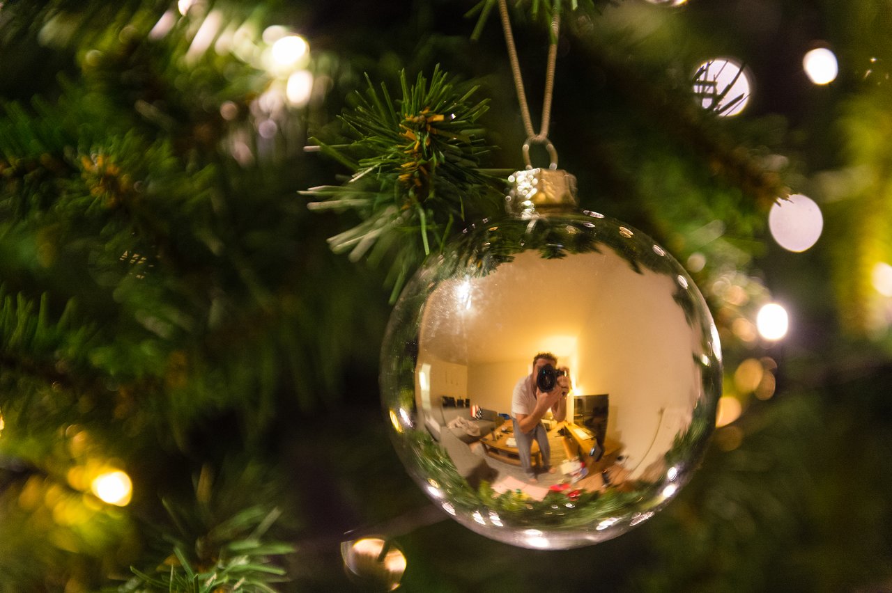 A photographer is reflected in a shiny Christmas ornament hanging on a decorated tree with warm lights.