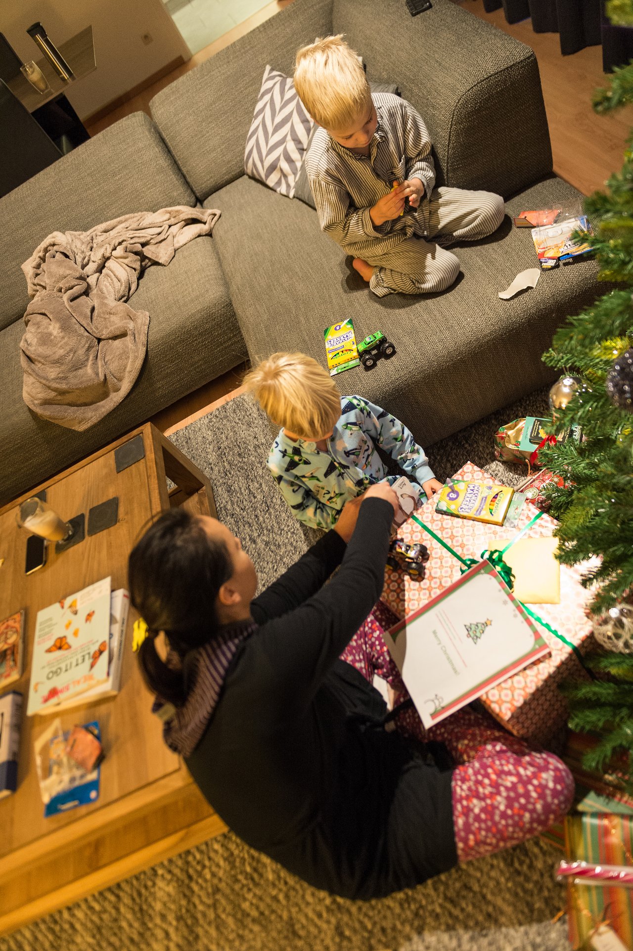 A woman and two children sit near a Christmas tree, unwrapping presents and playing with toys.