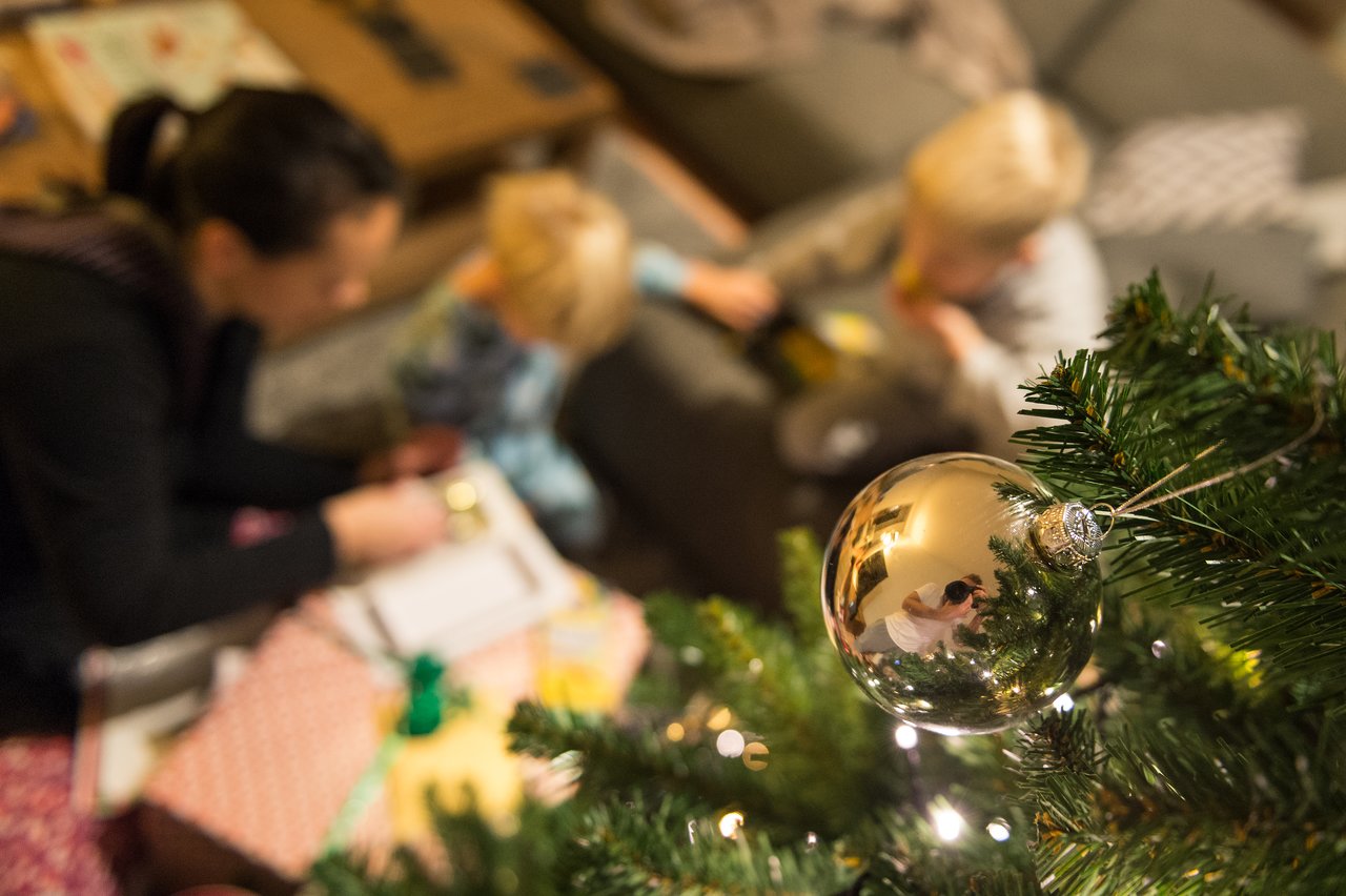 A reflective ornament on a Christmas tree captures a photographer, while a family wraps presents in the background.