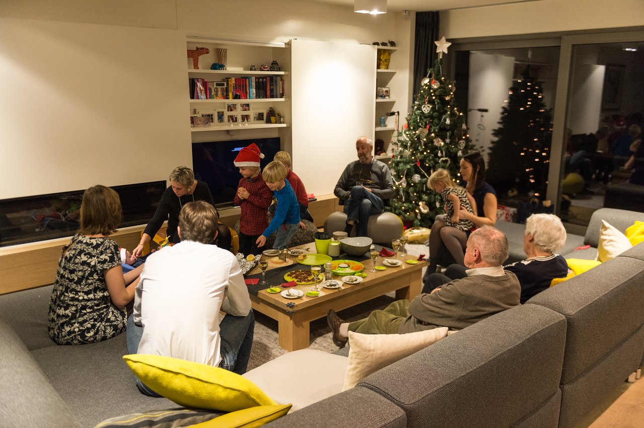 Family and friends gather in a living room, enjoying food and conversation near a decorated Christmas tree.