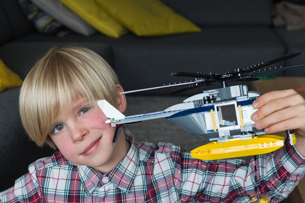 A child in a plaid shirt smiles while holding up a LEGO helicopter.
