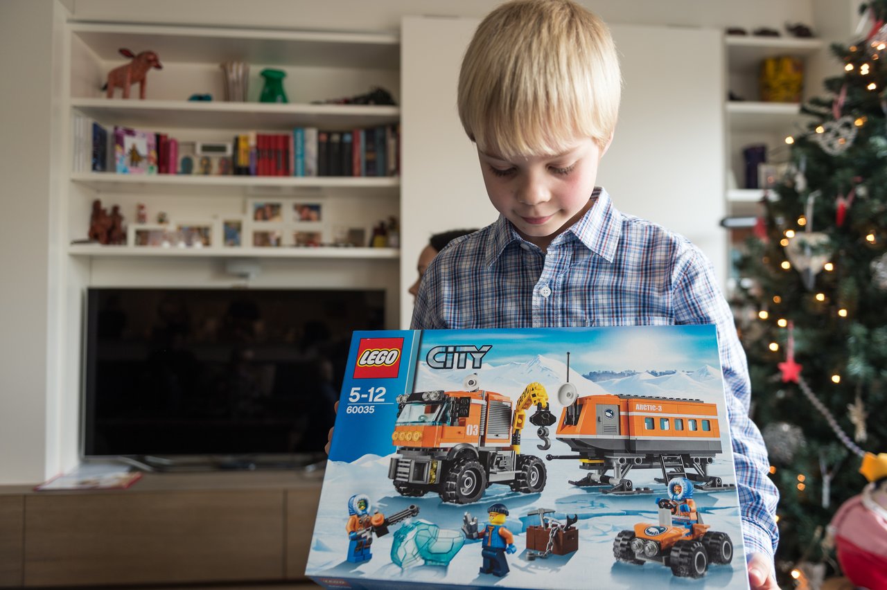 A young boy in a plaid shirt holds a LEGO City set, looking at it with excitement near a Christmas tree.
