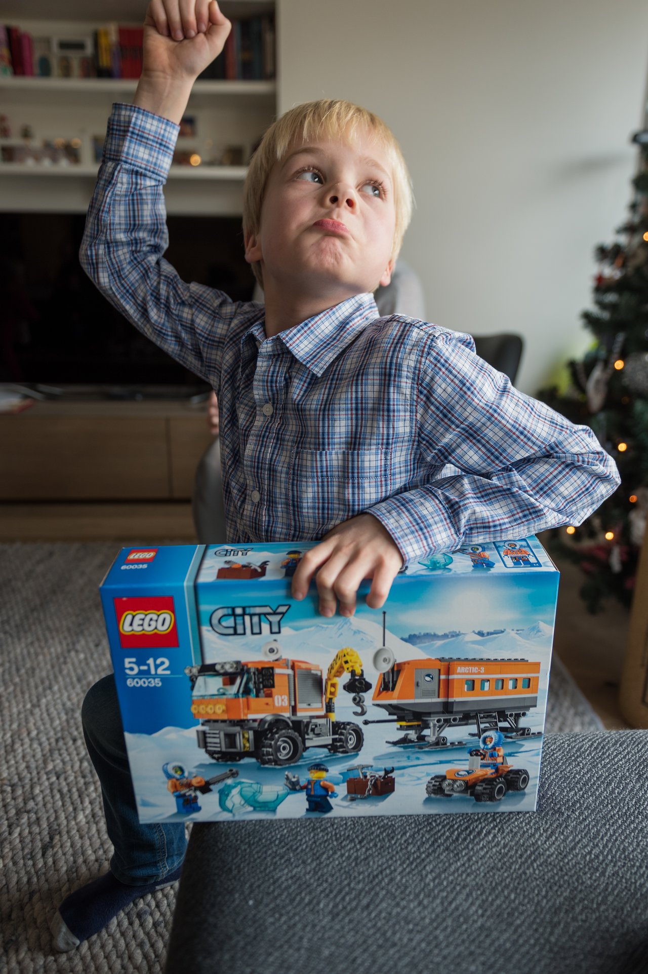 A young boy holds a LEGO City set while raising his arm, looking excited.
