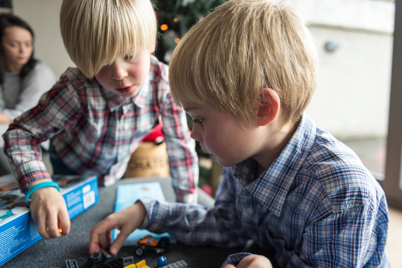 Two children in plaid shirts play with toy cars and building blocks near a Christmas tree.