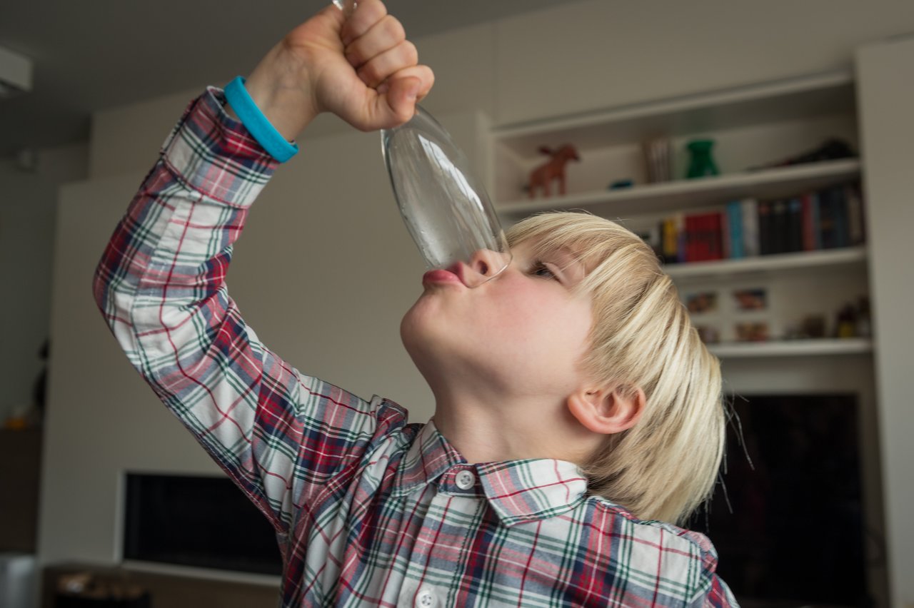 A young child in a plaid shirt tilts an empty glass upside down, pretending to drink from it.