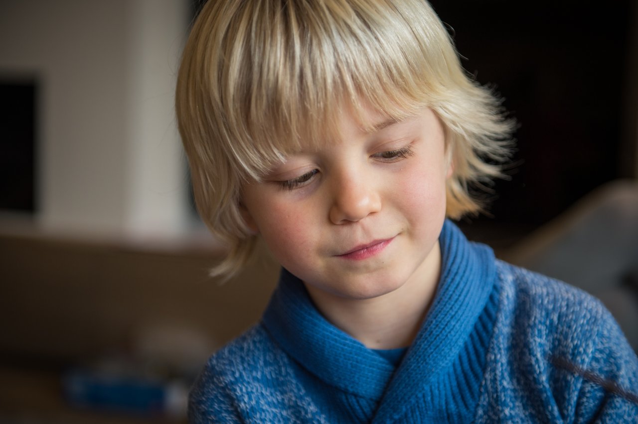 A young child in a blue sweater looks down with a calm expression indoors.