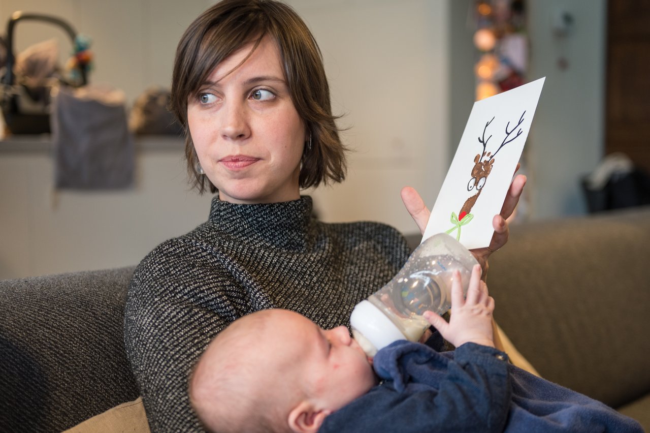 A woman holds a Christmas card with a reindeer drawing while feeding a baby with a bottle.