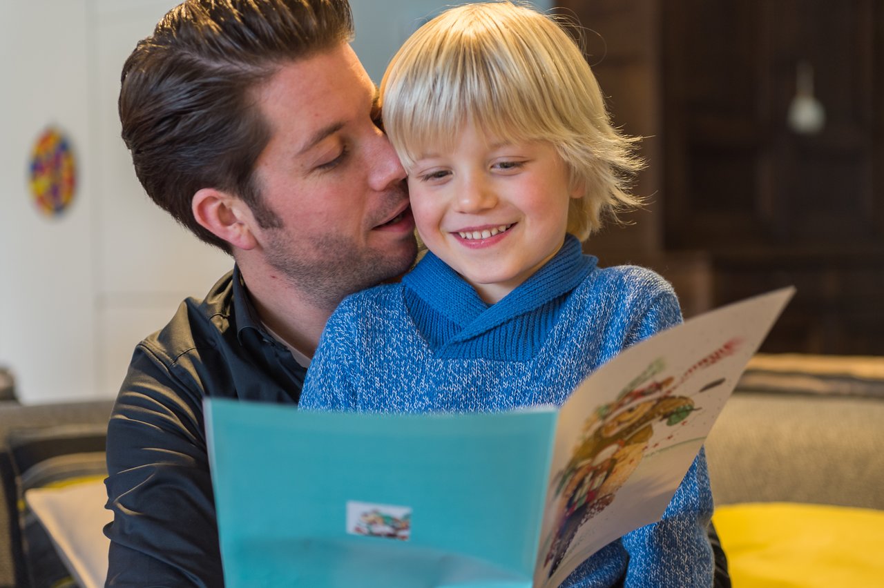 A smiling child in a blue sweater reads a Christmas card while sitting with an adult who is leaning in.