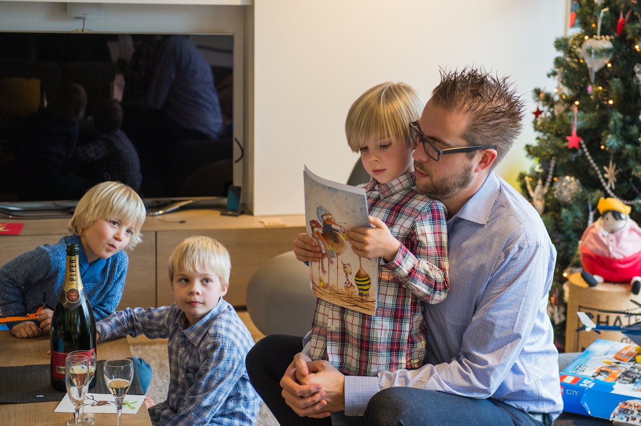 A man sits with a child reading a book, while two other children sit nearby.