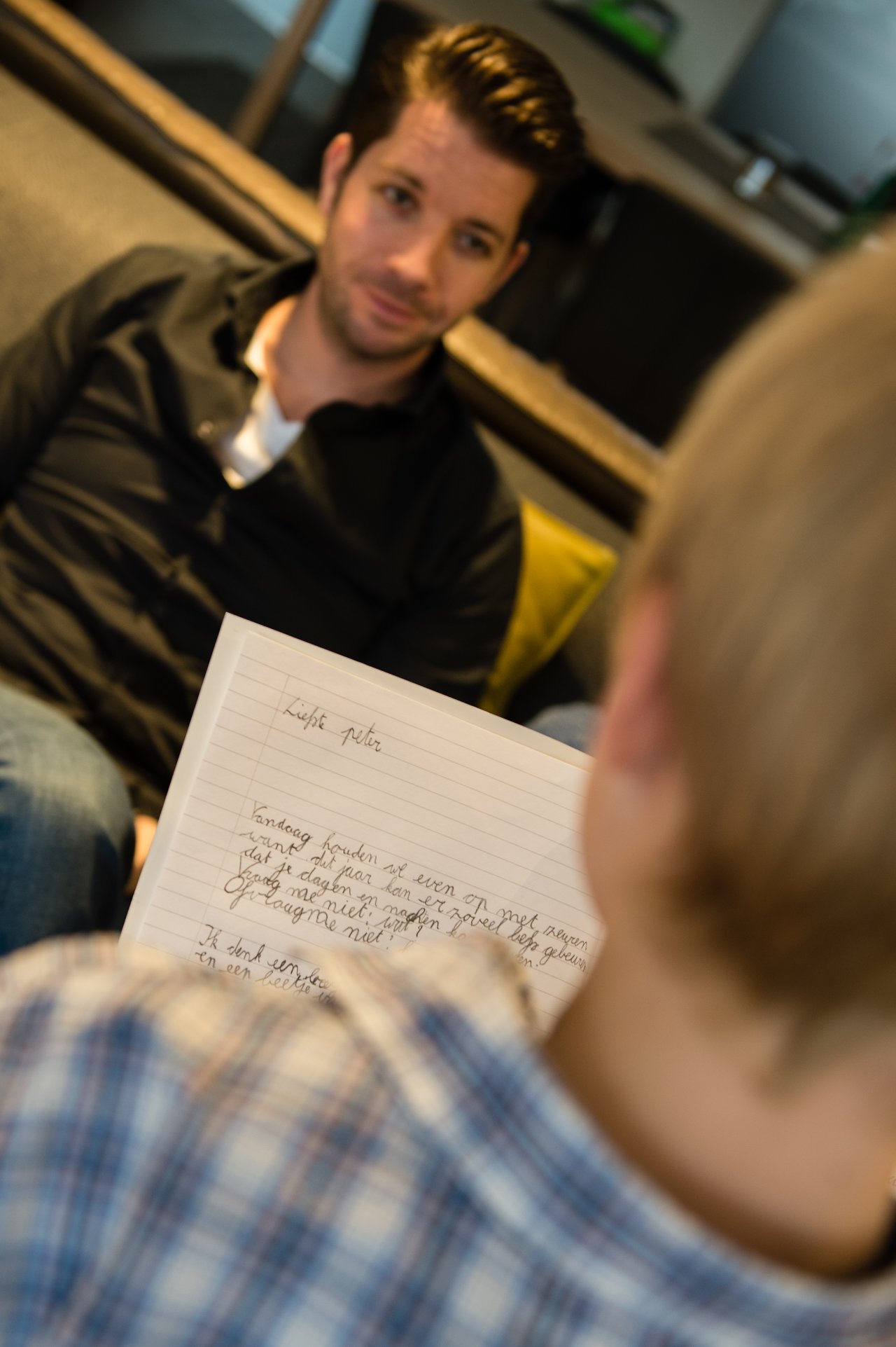 A person in a plaid shirt reads a handwritten letter aloud to someone sitting on a couch, listening attentively.