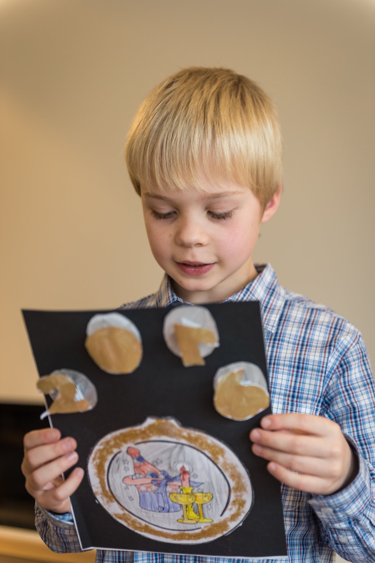A young boy in a plaid shirt holds up a handmade Christmas craft with drawings and cut-out shapes.