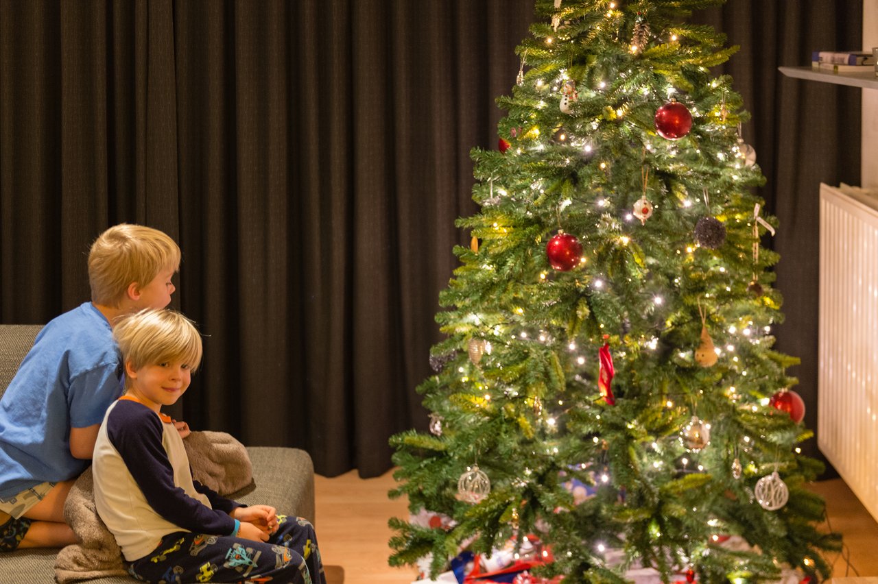 Two children sit on a couch, smiling and looking at a decorated Christmas tree with glowing lights.
