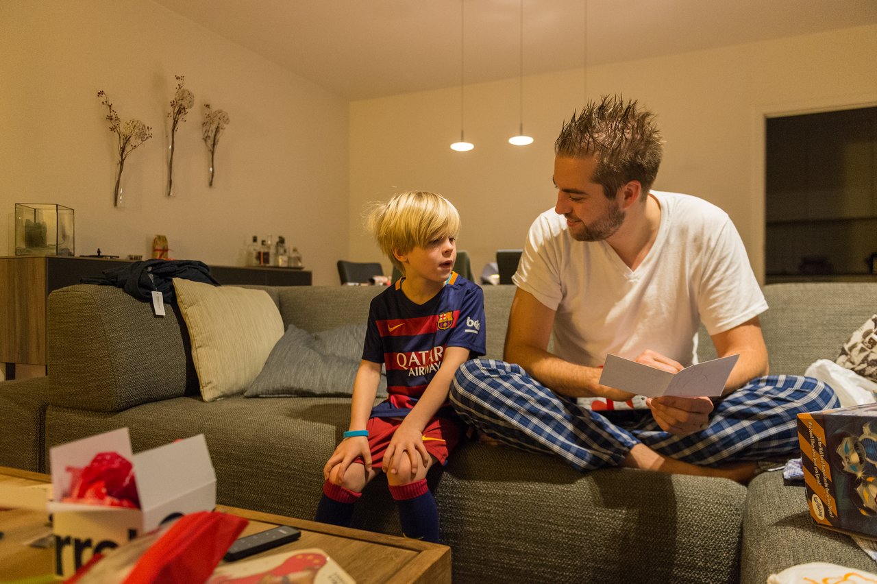 A man in pajamas sits on a couch, reading a card while talking to a young boy in a soccer jersey.