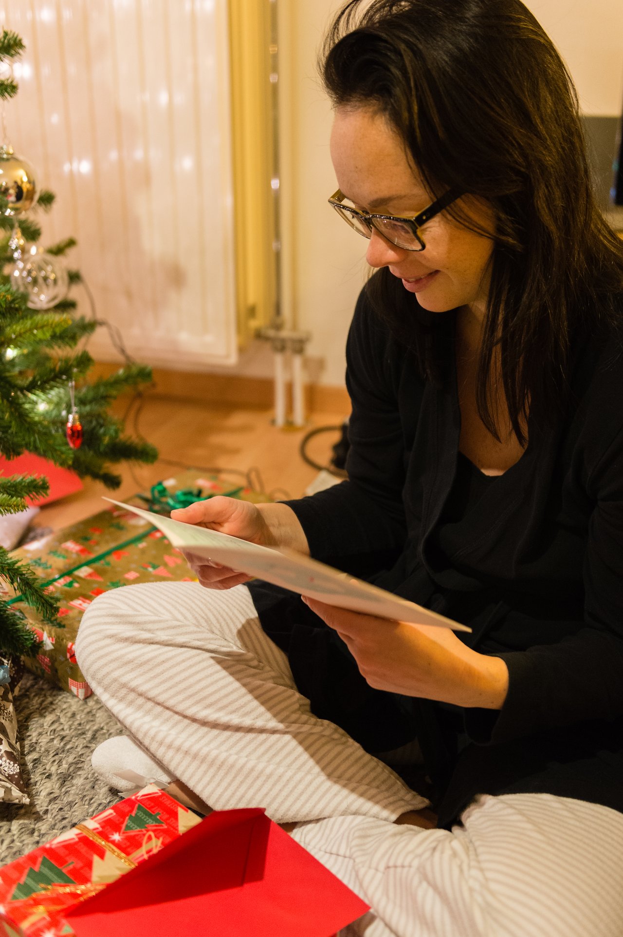 A woman sits by a Christmas tree, smiling while reading a card with presents around her.