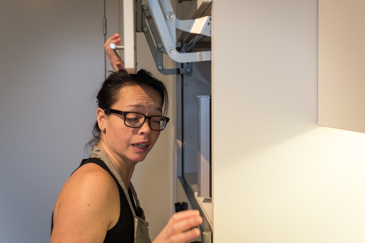 A woman wearing glasses and an apron stands in a kitchen, reaching up to adjust an overhead cabinet.