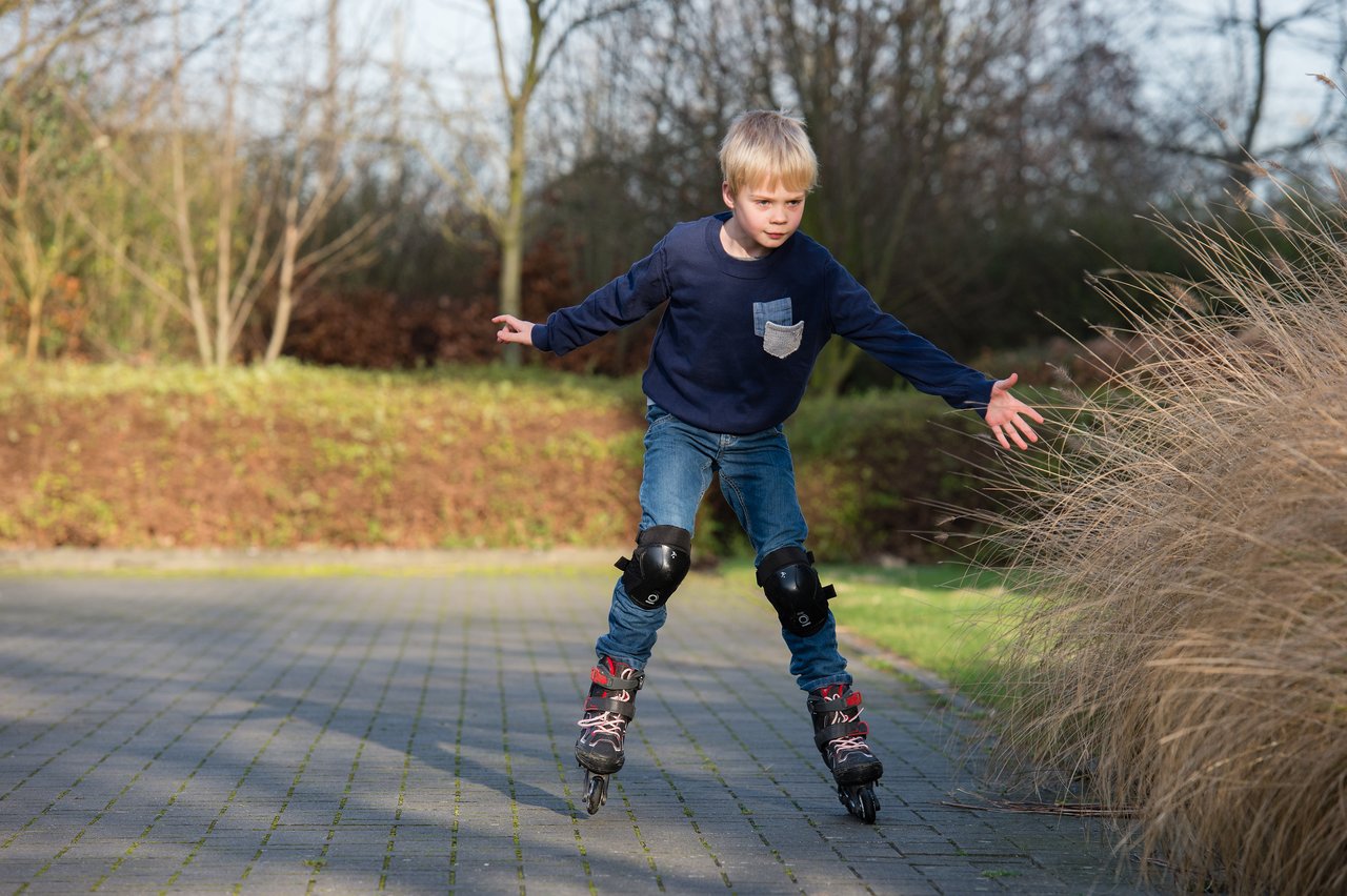 A young boy wearing knee pads rollerblades on a paved path, balancing with his arms outstretched.