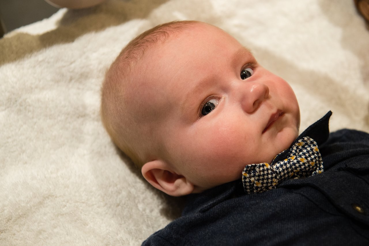 A baby wearing a bow tie and dark outfit lies on a soft surface, looking slightly to the side.