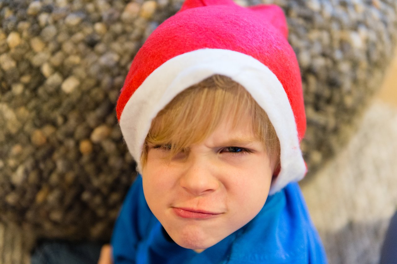 A child wearing a Santa hat and blue shirt makes a playful, scrunched-up face while looking at the camera.
