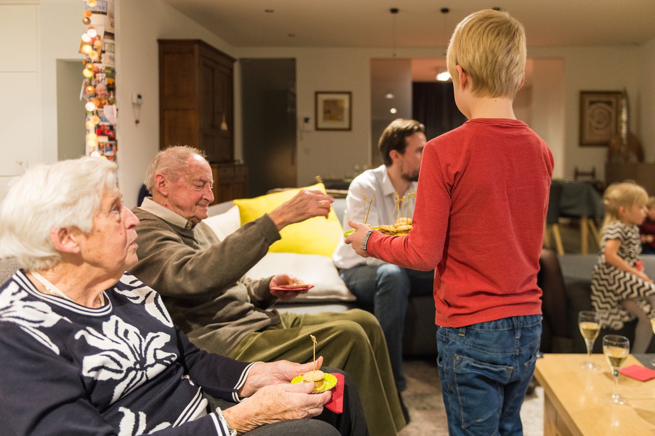 A young boy in a red shirt serves snacks to an elderly man sitting on a couch during Christmas.
