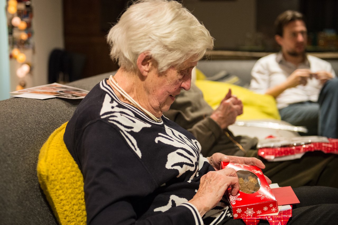 An elderly woman sits on a couch, opening a red Christmas gift box with cookies inside.