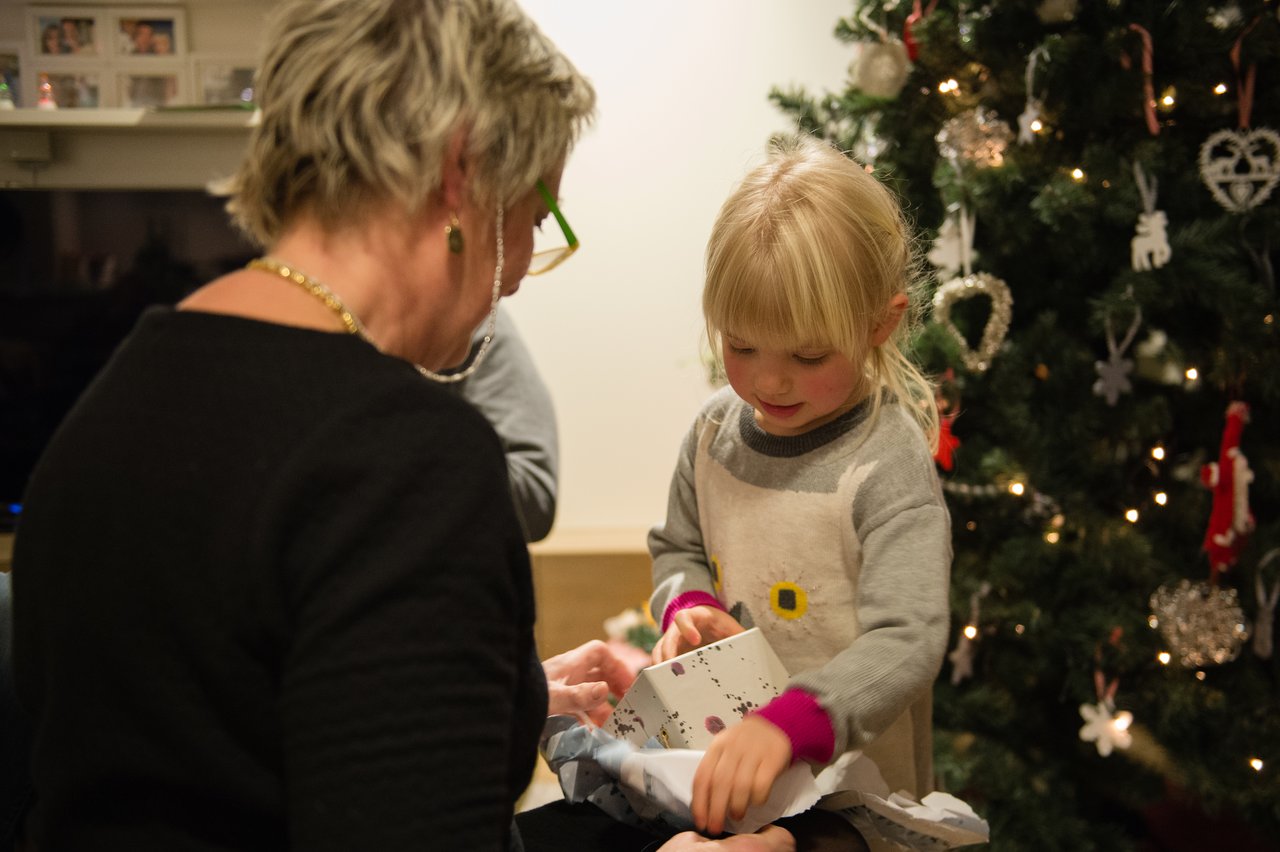 A young girl excitedly opens a Christmas present beside a decorated tree, while an older woman watches nearby.