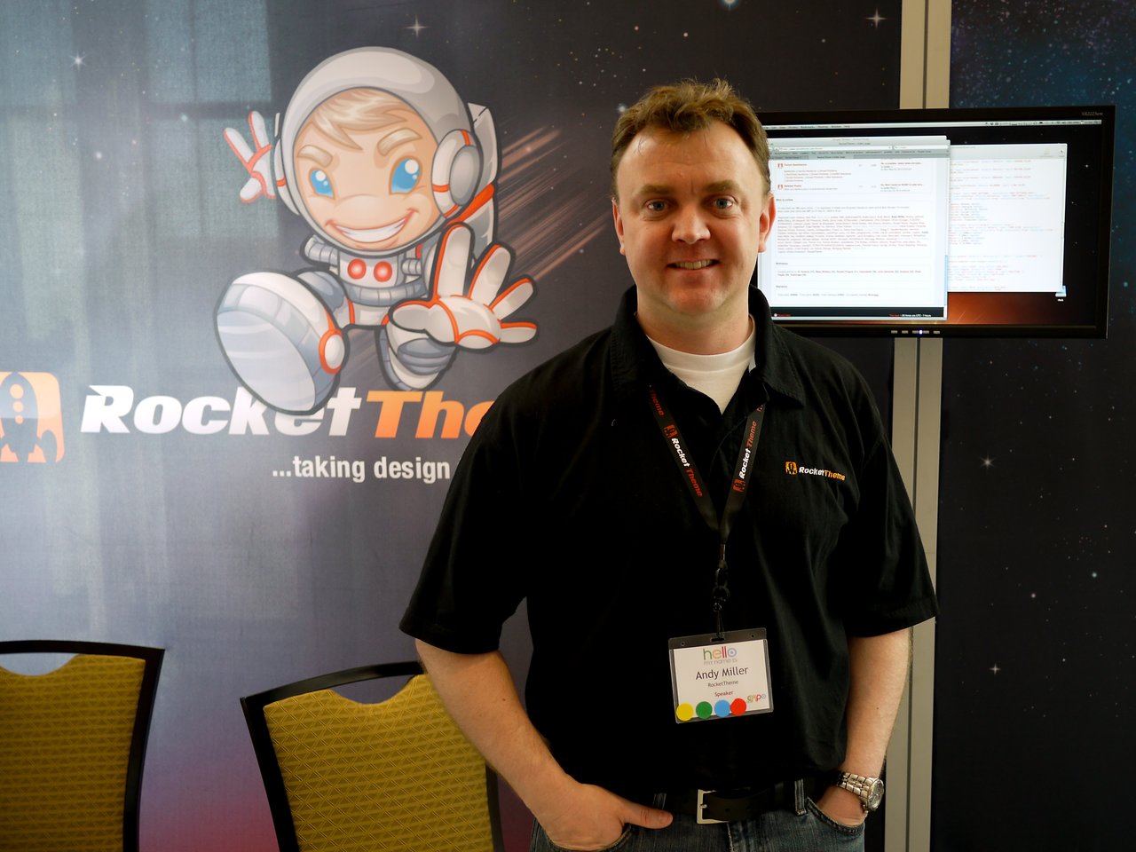 A man wearing a RocketTheme shirt stands in front of a RocketTheme booth with a screen displaying code.