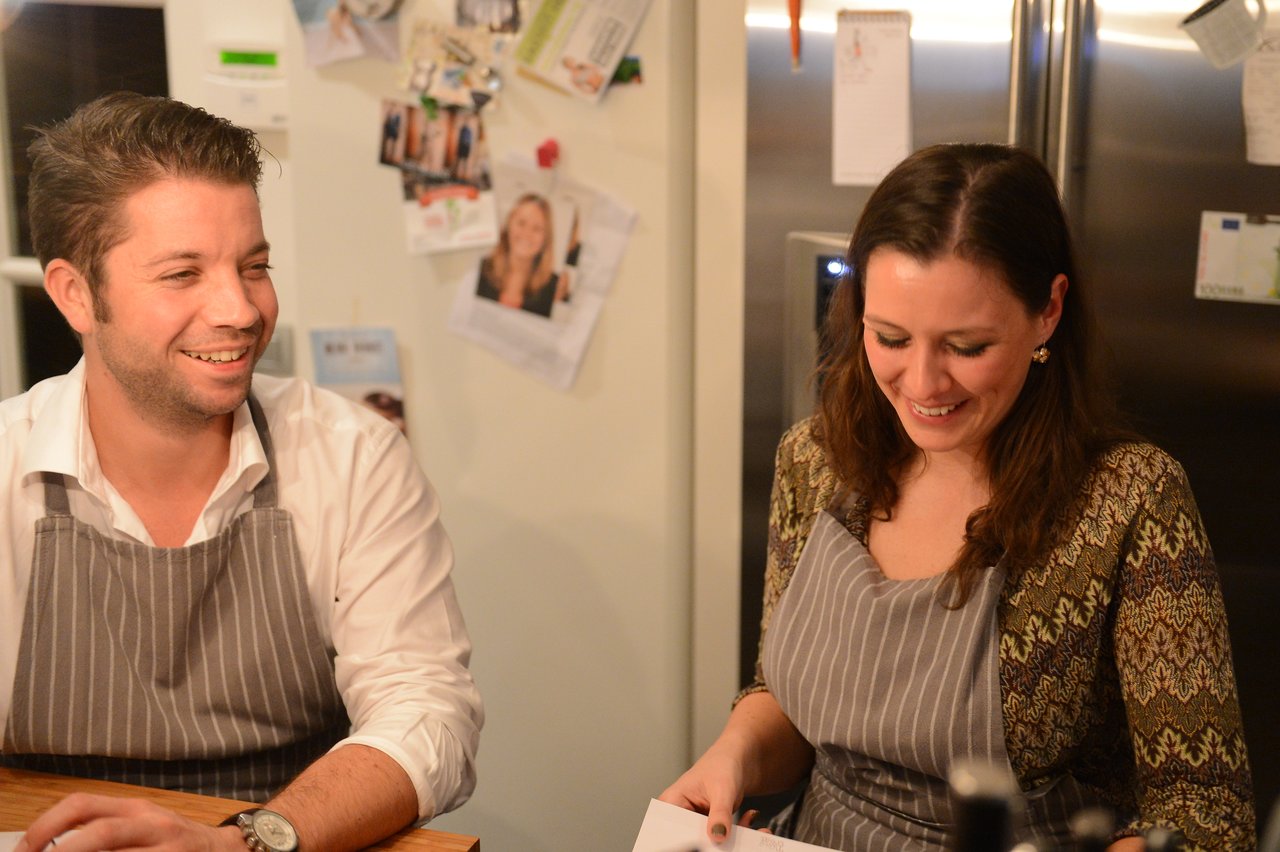 Two people wearing aprons smile while participating in a cooking class, looking at a piece of paper.