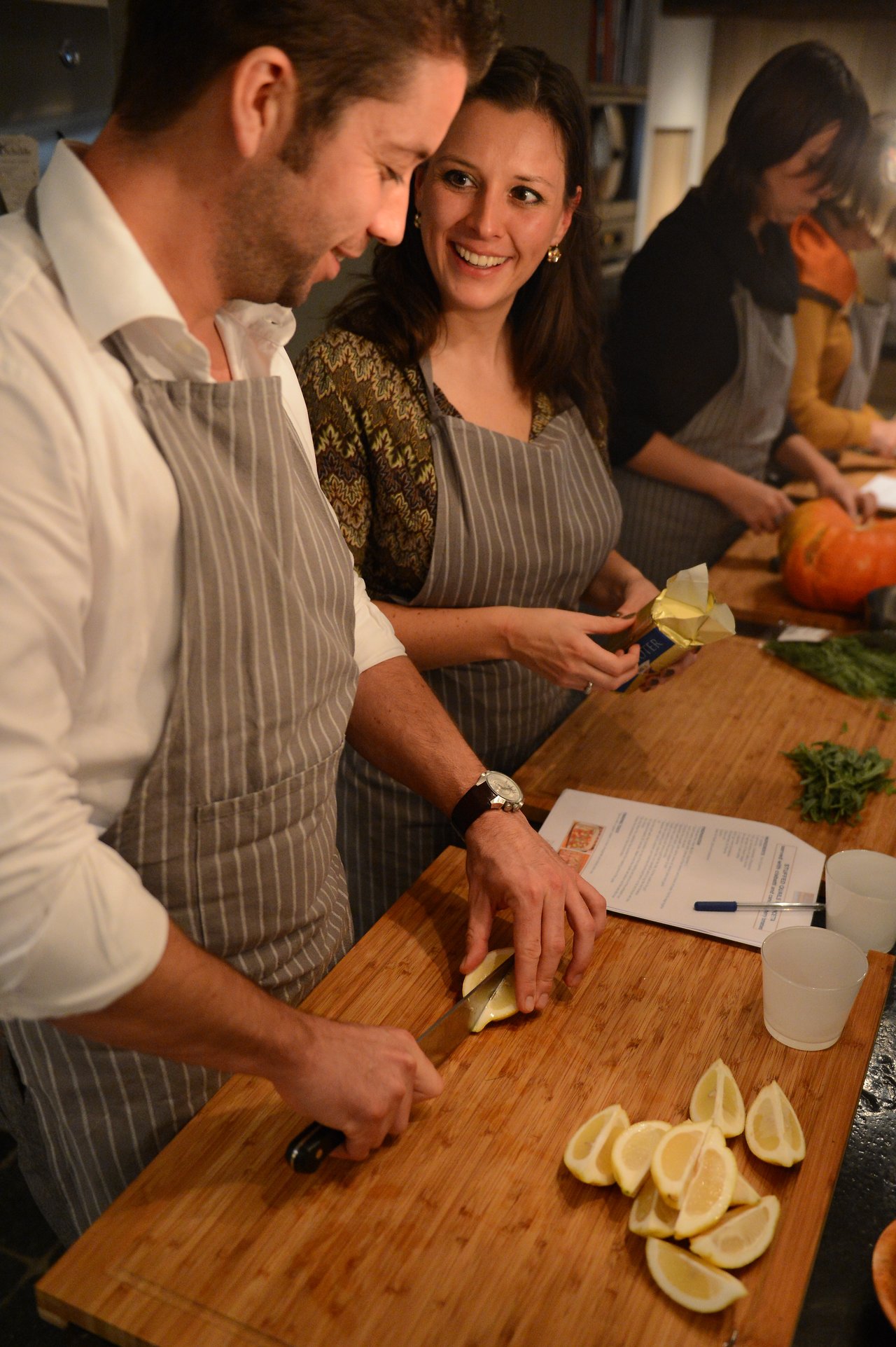 Two people wearing aprons prepare ingredients together, slicing lemons and butter during a cooking class.
