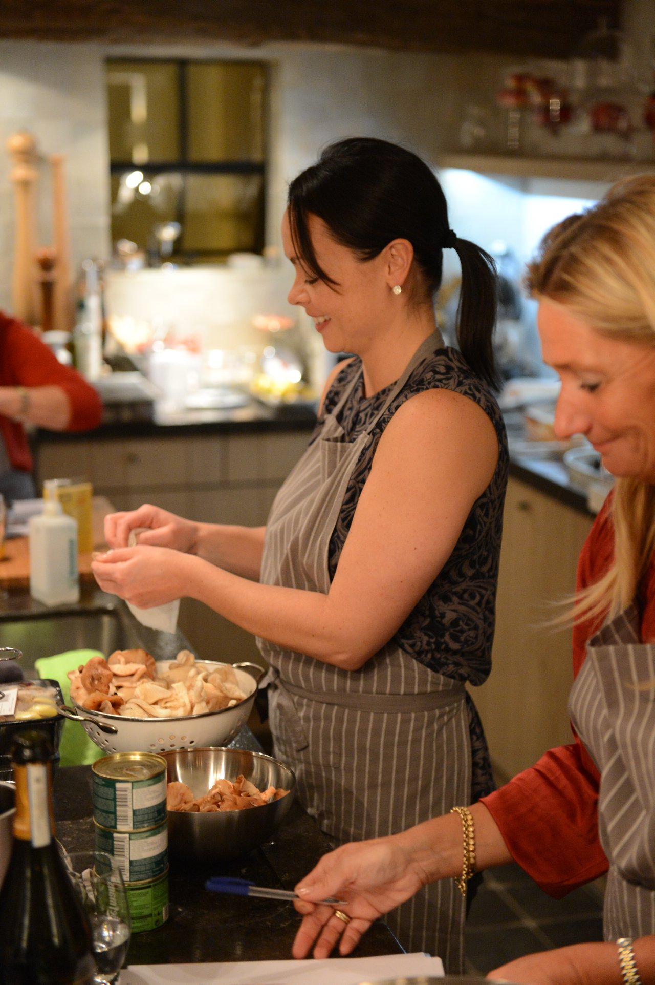 Two people wearing aprons prepare ingredients in a kitchen, with one peeling or handling food over a bowl.