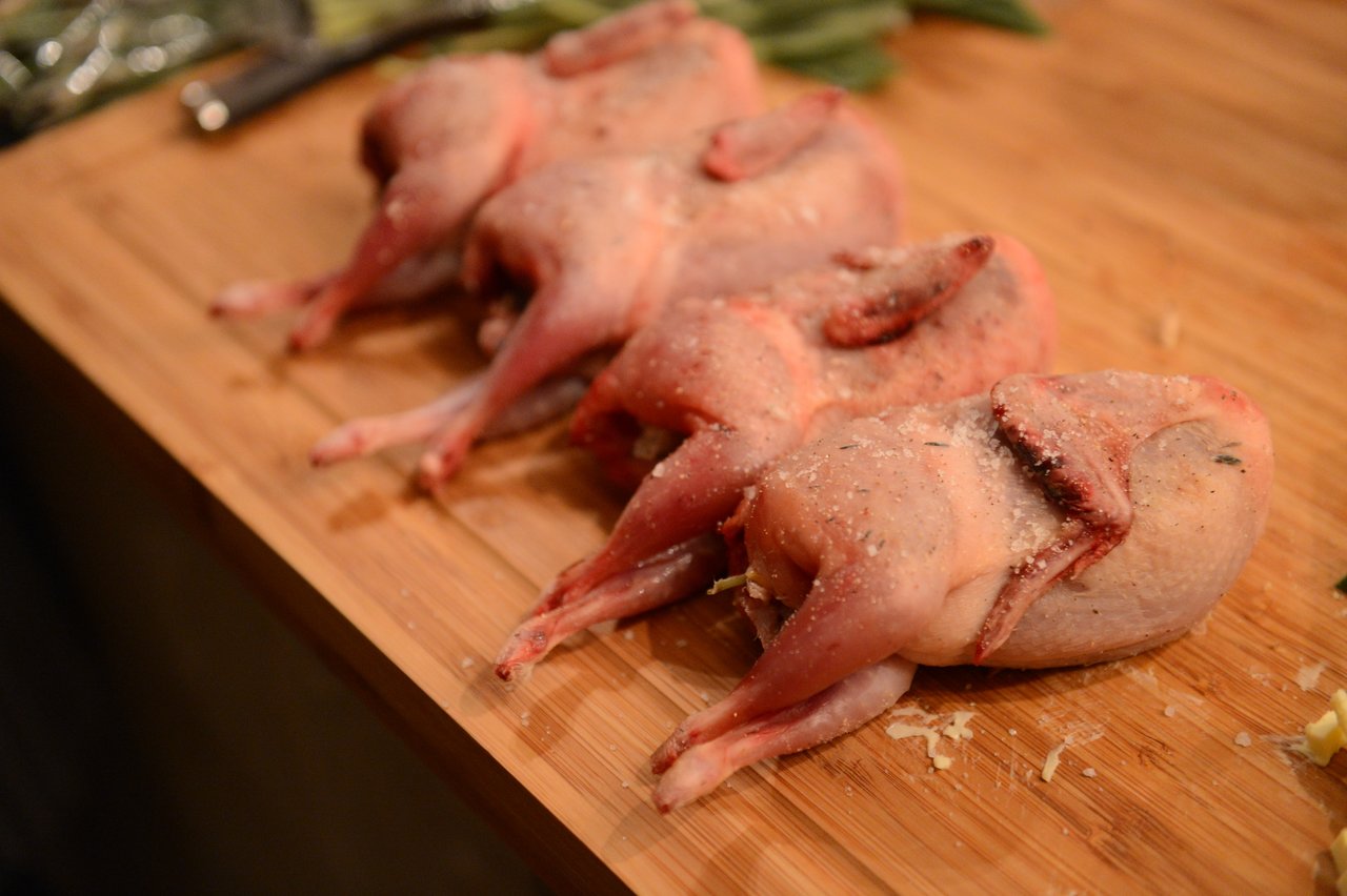 Four raw, seasoned quails are arranged on a wooden cutting board, prepared for cooking.