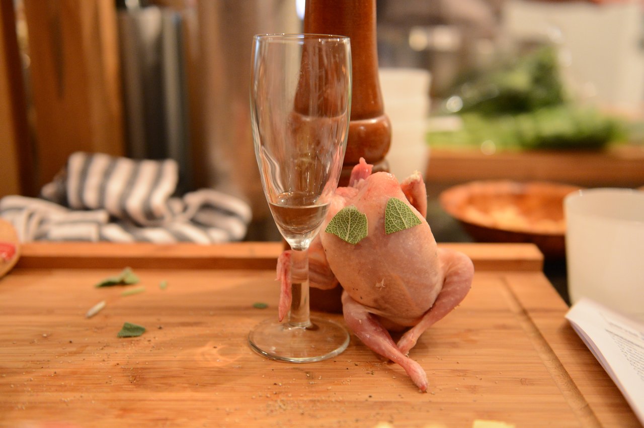 A raw, plucked quail is posed upright against a glass with sage leaves on its back, on a cutting board.
