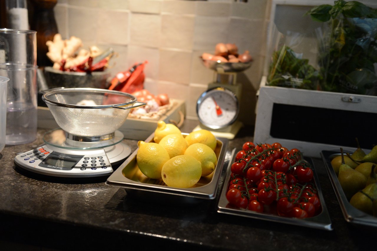 Fresh lemons, cherry tomatoes, and pears on trays beside a kitchen scale and strainer during a cooking class.