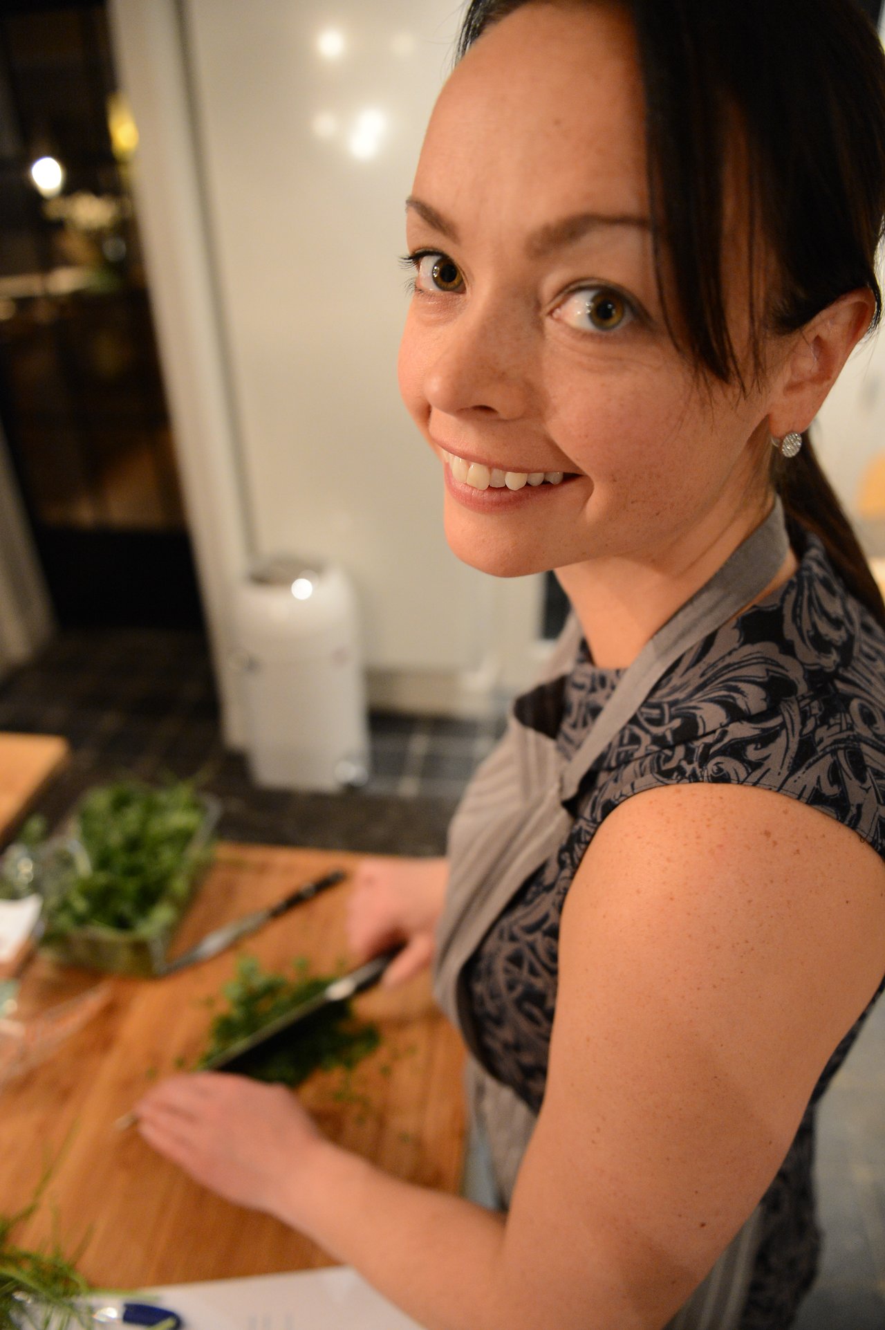 A woman in an apron smiles while chopping fresh herbs on a wooden cutting board during a cooking class.